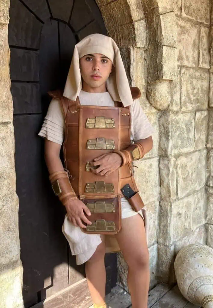 Homem vestido como gladiador romano, com armadura de couro e acessórios no estilo antigo, posando ao lado de uma parede de pedra, em cenário de temática histórica ou medieval.