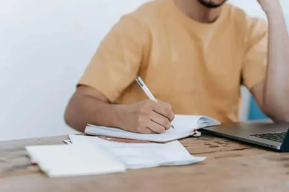 Estudante estudando e fazendo anotações, com laptops e papéis em uma mesa, buscando atualização legal e jurídica.