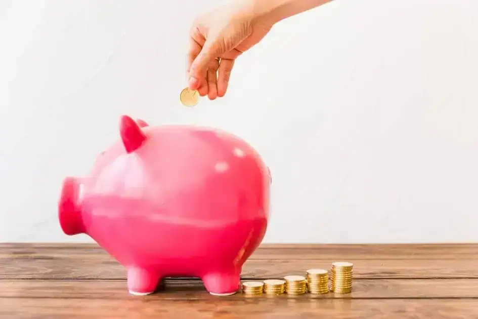Dinheiro sendo colocado em um cofrinho rosa, simbolizando economia ou investimento, com pilhas de moedas ao redor, conceito de finanças, economia doméstica e planejamento financeiro, relacionado a notícias de direito e economia.