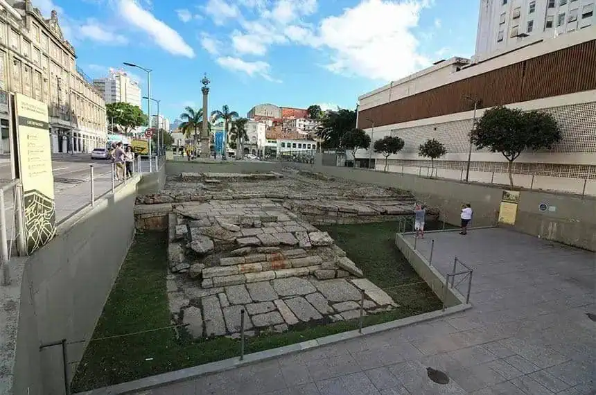 Muros antigos em ruínas no centro de uma cidade brasileira, com pessoas ao redor e edifícios modernos ao fundo, representando conflito entre história e urbanização.