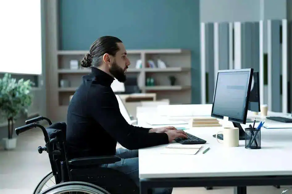 Homem em cadeira de rodas trabalhando em escritório com computador, acessibilidade e inclusão no ambiente de trabalho, direito às facilidades para pessoas com deficiência, direito hoje notícias.
