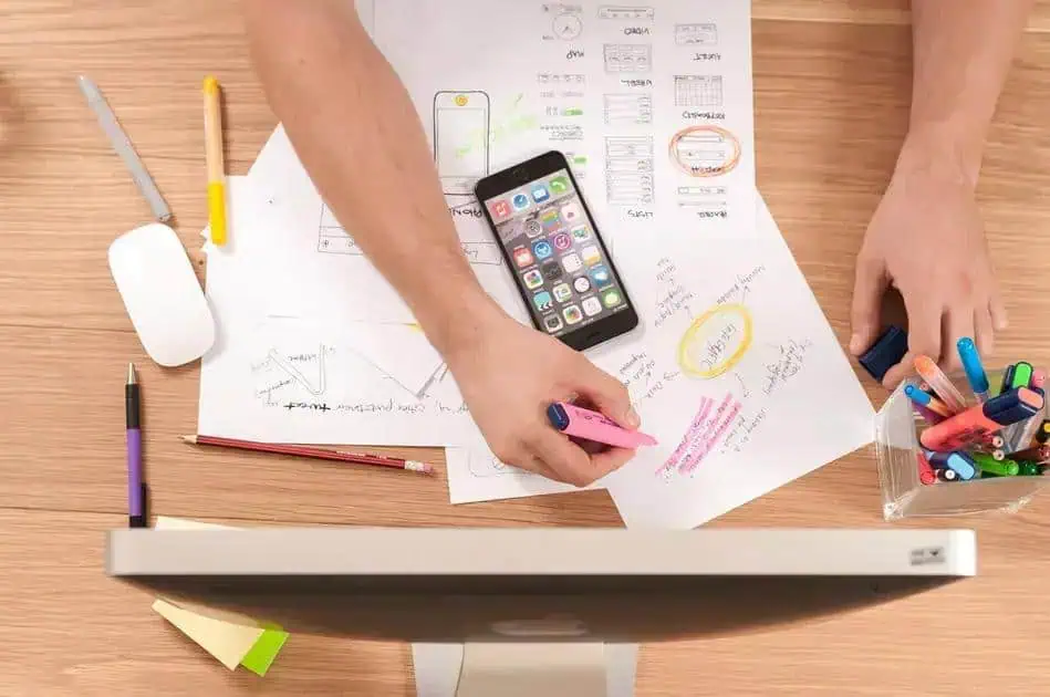 Desorganização na mesa de trabalho com documentos, canetas coloridas, smartphone e computador, representando trabalho, estudos e organização de tarefas.