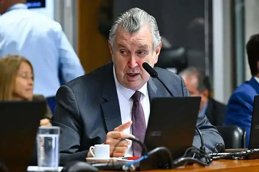 A3. Homem de terno falando em comissão durante reunião do tribunal, ambiente formal, com pessoas ao fundo, computadores e microfones, foco em debate jurídico e notícias de direito.