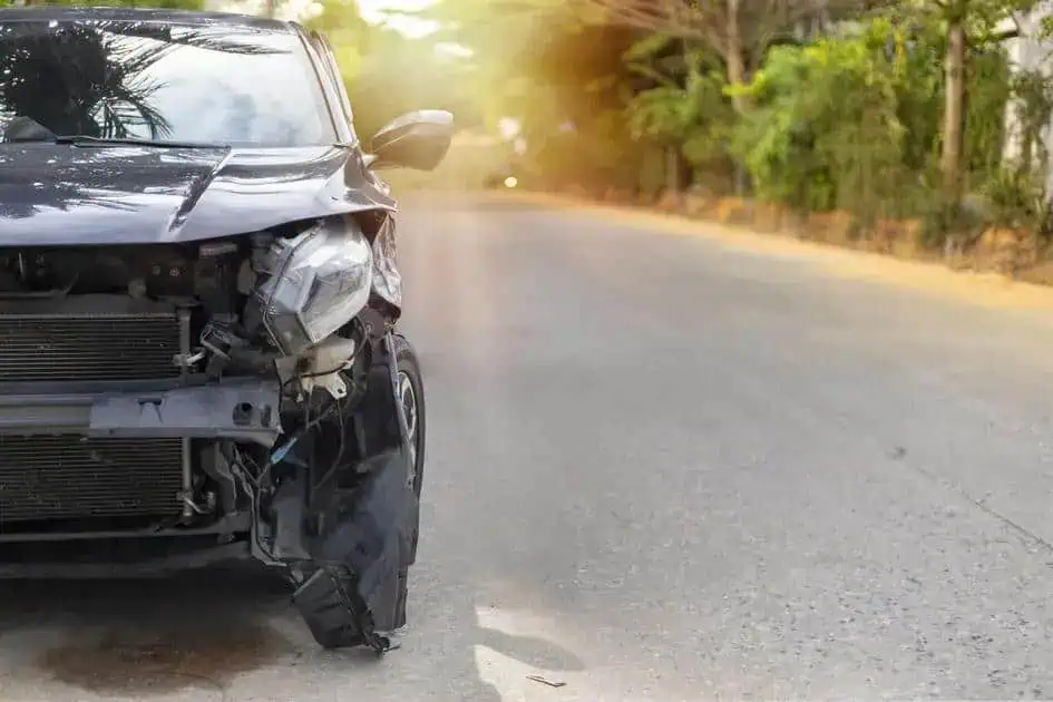 Carro com colisão frontal em estrada de asfalto, com danos visíveis na parte dianteira, refletindo acidente de trânsito. Fundo de árvores e iluminação natural, foco na importância de segurança nas vias.