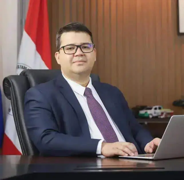 Homem de terno e gravata sentado na mesa de escritório, com laptop e bandeira do Brasil ao fundo, representando notícias de direito e política.