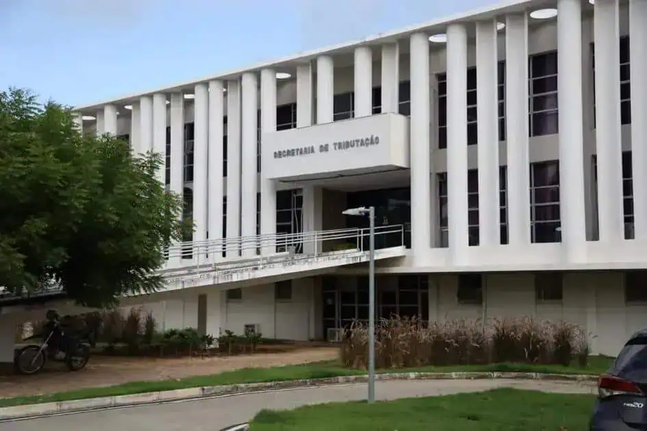 Escritório da Secretaria de Tributação, prédio moderno com colunas largas, fachada branca e janelas altas, localizado em área urbana com árvores, veículos e iluminação pública ao redor, em destaque na área de Direito e Tributação.