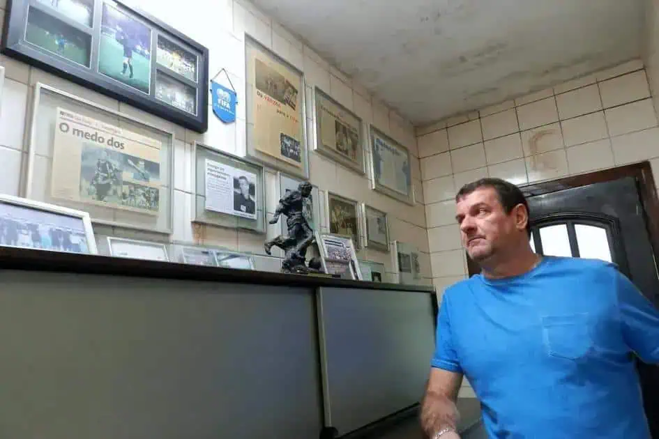 O homem observando uma parede decorada com diversos quadros, fotografias e uma escultura, em ambiente que remete a uma sala de notícias ou escritório de direito.