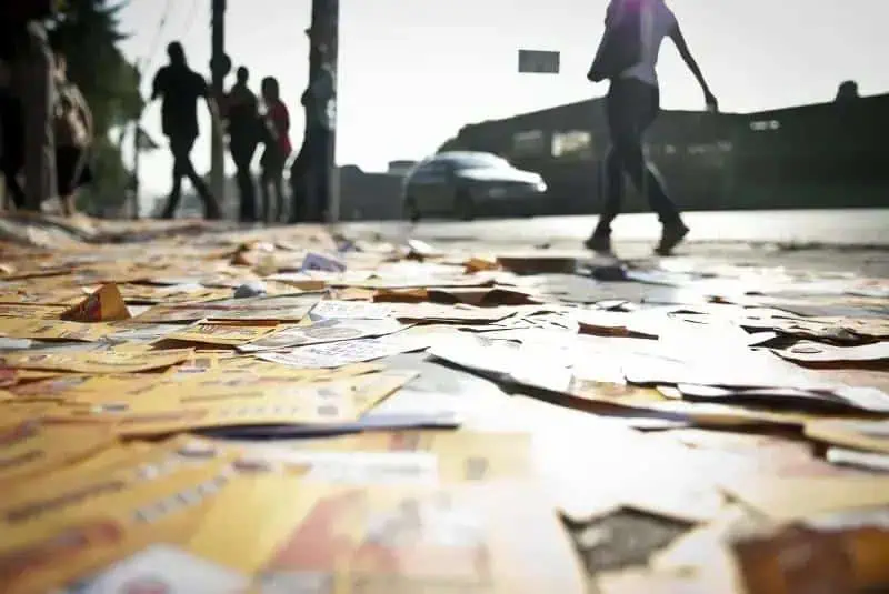 Pilhas de jornais e documentos espalhados no chão em uma rua movimentada, pessoas ao fundo, cenário urbano, iluminação de fim de tarde, símbolo de notícias e informação, referente a direito, jornalismo e atualidades no Brasil.