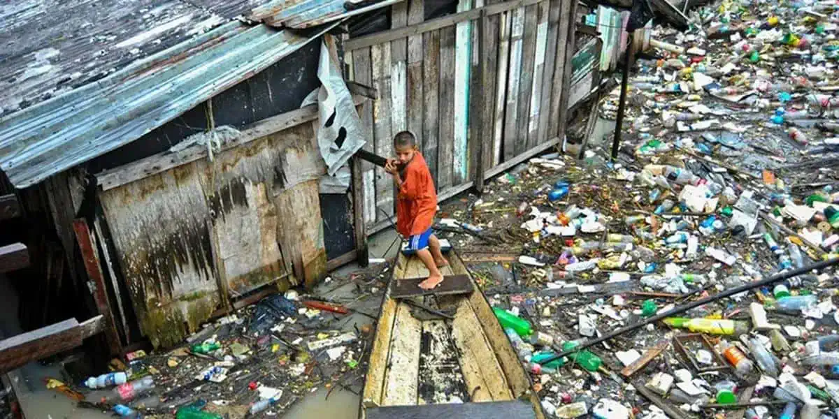 Menino atravessando uma ponte improvisada sobre área de lixo, situação de pobreza e condições precárias de moradia.
