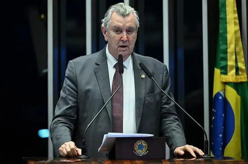 Senador brasileiro falando em sessão do Congresso com bandeira do Brasil ao fundo.