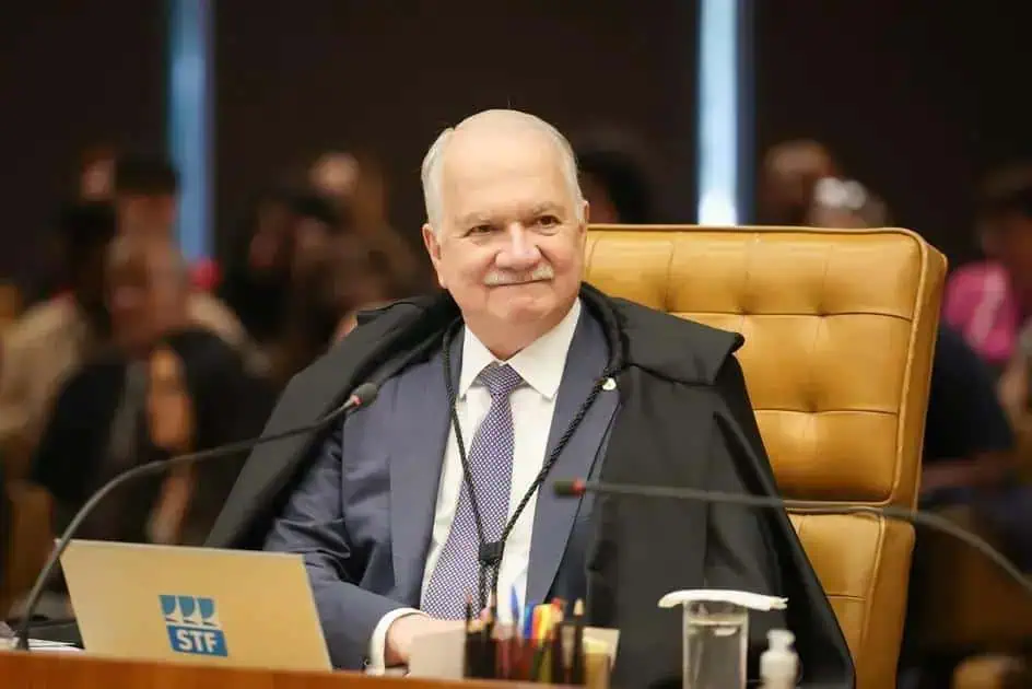 Homem idoso, juiz ou autoridade jurídica, sentado em tribunal, usando toga e vestimenta formal, com pessoas ao fundo, simbolizando ambiente de julgamento ou sessão de tribunal, Direito Hoje Notícias, foco em justiça e direito.