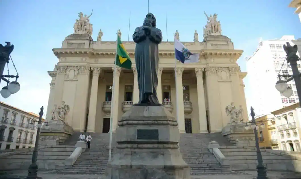 Estátua em frente ao Palácio da Justiça em São Paulo, símbolo do direito e da justiça no Brasil, com bandeiras nacionais e estaduais ao fundo, destaque para notícias e atualidades jurídicas.