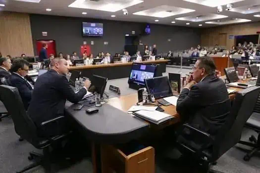 Audiência judicial com advogados e profissionais do direito em ampla sala de tribunal, com computadores e documentos discutindo casos legais e legislativos.