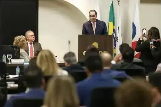Homem em palestra ou evento jurídico, com bandeiras no fundo, falando ao público em ambiente formal.