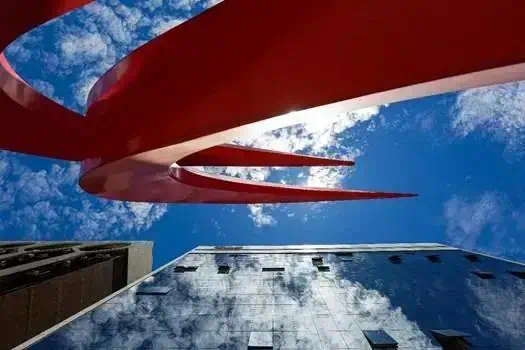 Escultura moderna vermelha em edifício alto sob céu claro com nuvens, destaque em notícias de direito e cultura urbana.