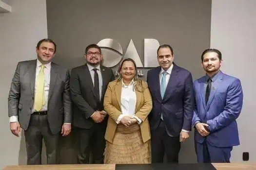 Homens e mulher de negócios posando juntos em ambiente profissional em frente a logo da entidade.