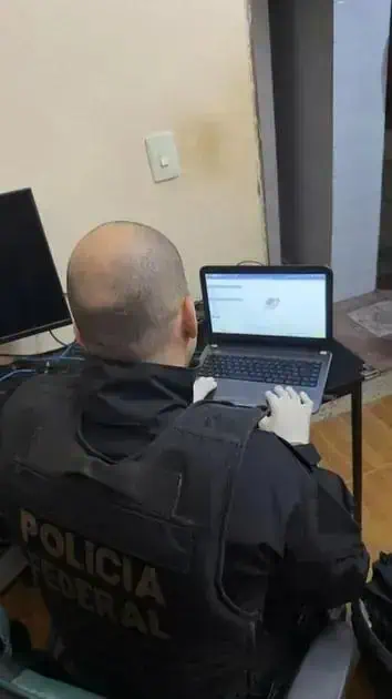 Policial da Polícia Federal usando laptop para investigação digital em ambiente interno, concentrado na análise de dados.