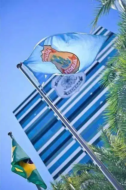 Bandeira do Brasil e da Polícia Federal ao vento, com fundo de arranha-céu e árvores verdes, simbolizando a presença e atuação do direito na sociedade brasileira.