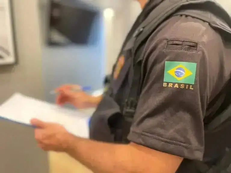 Policial militar com uniforme do Brasil, segurando documentos, em ambiente de trabalho.