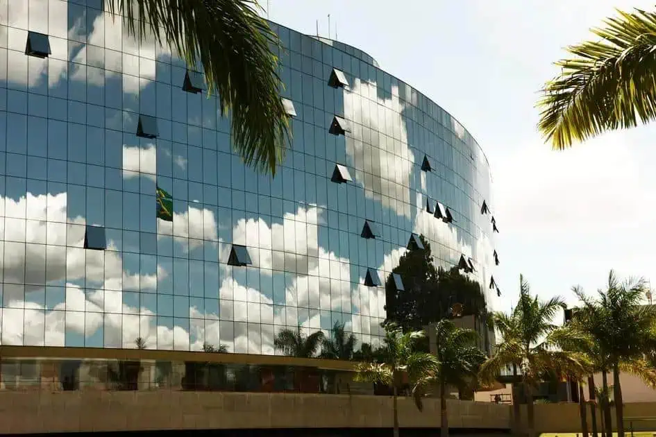 Edifício moderno com fachada de vidro refletindo o céu e nuvens, cercado por palmeiras, representando destaque em notícias de direito, negócios e notícias jurídicas no Brasil.