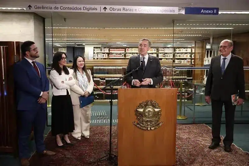 Deputado em pronunciamento na biblioteca do congresso com grupo de colegas ao fundo, ambiente formal com livro e gravatas, destaque nas notícias de direito hoje, evento político ou jurídico relevante.