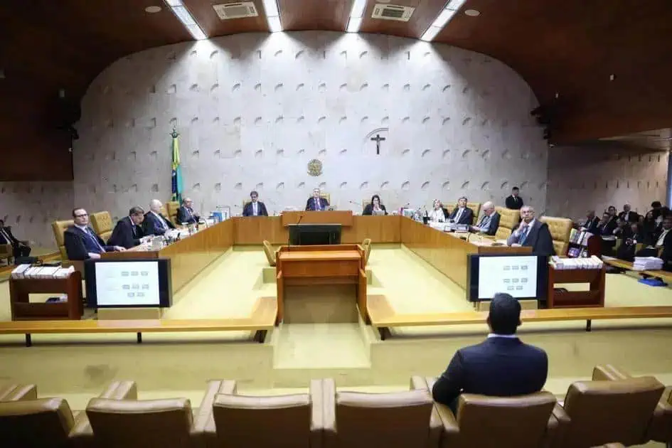 Pessoas sentadas em reunião do tribunal, discutindo temas jurídicos e sobre direito, em ambiente formal e oficial, na sala de audiência do judiciário brasileiro, com destaque para a bandeira e símbolos nacionais.