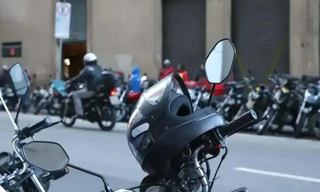 Capacete de moto estacionado na rua com vários motociclistas ao fundo, em ambiente urbano, destacando segurança no trânsito para artigos de direito e leis de trânsito.