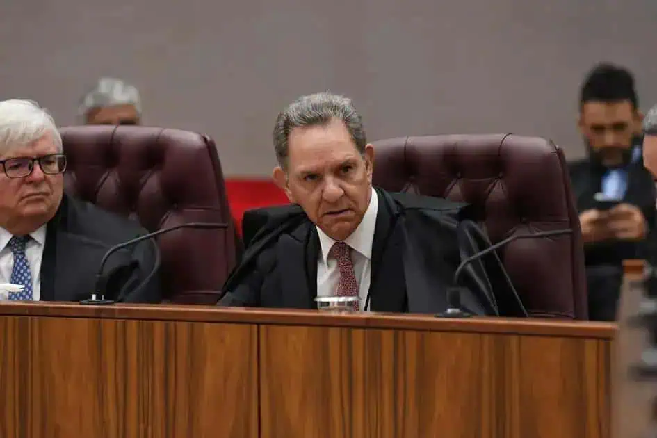 Juiz do tribunal durante sessão judicial com profissionais do direito e magistrados ao fundo.
