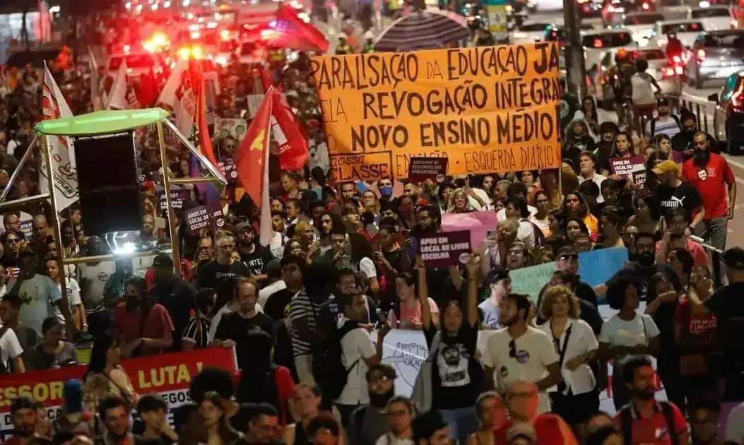 Discurso de protesto exige melhorias na educação pública, com cartazes e faixas pedindo revogação de medidas que prejudicam o ensino médio, destacando mobilização de estudantes e apoiadores por direitos educacionais.