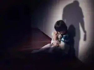 Menina triste e solitária sentada no chão com sombra de uma figura ameaçadora ao fundo, simbolizando vulnerabilidade e risco para crianças.
