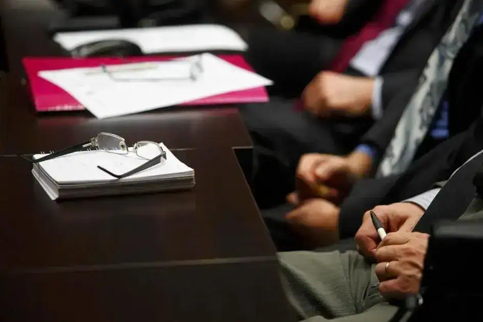Somentes uma mesa de reunião com documentos, óculos e pessoas de terno presentes em uma audiência ou reunião jurídica.