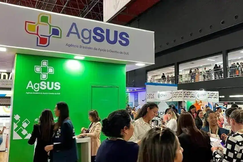 AgSUS, stand de apoio à gestão do SUS, em evento de saúde pública, com muitas pessoas interagindo, refletindo a importância da saúde coletiva e profissionais de saúde em destaque no setor de assistência social.