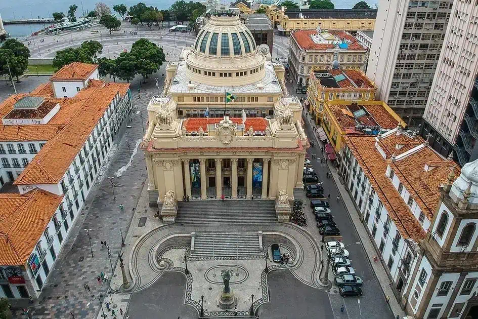 Palácio dos Correios em uma vista aérea em São Paulo, destacando sua arquitetura neoclássica, telhado de vidro e a bandeira brasileira hasteada na fachada, cercado por edifícios históricos e área movimentada da cidade.