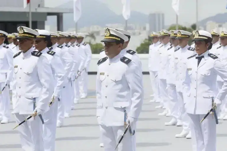 Formação de oficiais militares vestidos de uniformes brancos durante cerimônia oficial no topo de um edifício com vista urbana ao fundo.