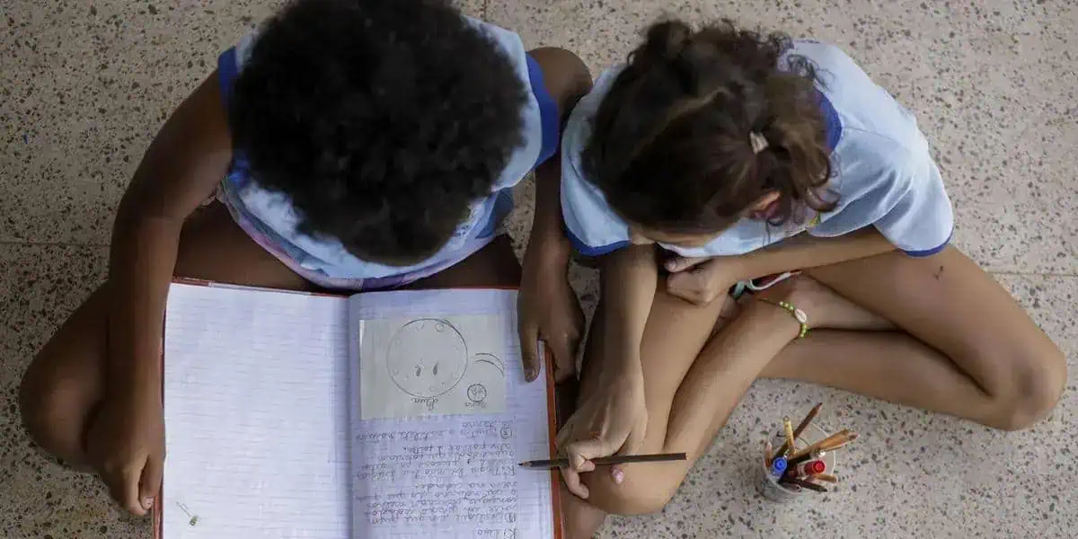 Crianças estudando juntos no chão com caderno e canetas, promovendo educação e inclusão social.