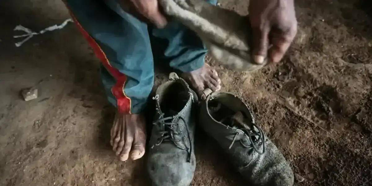 Sapatos velhos e usados em solo de terra, mãos sujas de terra e pés descalços, evidenciando trabalho rude ou situação de vulnerabilidade social, tema relevante para notícias de direitos e justiça.