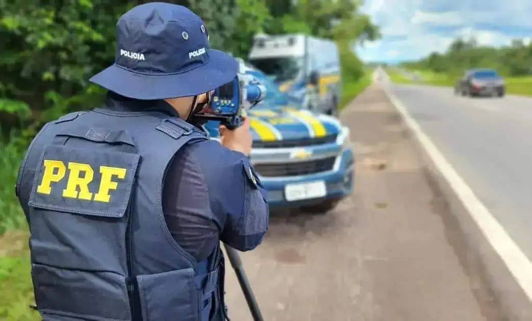 Policial da PRF realizando fiscalização de trânsito na rodovia com viaturas e veículos em background, promovendo segurança nas estradas brasileiras.