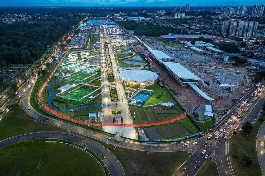 Praça de eventos e lazer com áreas verdes, pistas de caminhada, esportes e espaço para convivência na cidade ao entardecer, iluminada com teto de luzes e construções modernas em fundo urbano.