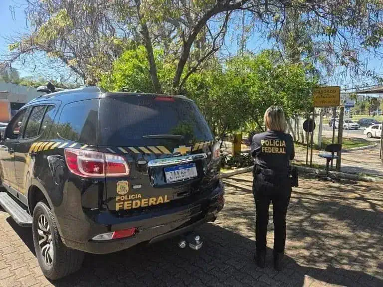 Polícia Federal em ação de fiscalização com carro oficial na rua, polícia feminina de costas observando a operação na área urbana sob céu claro, destaque em segurança e investigação policial.