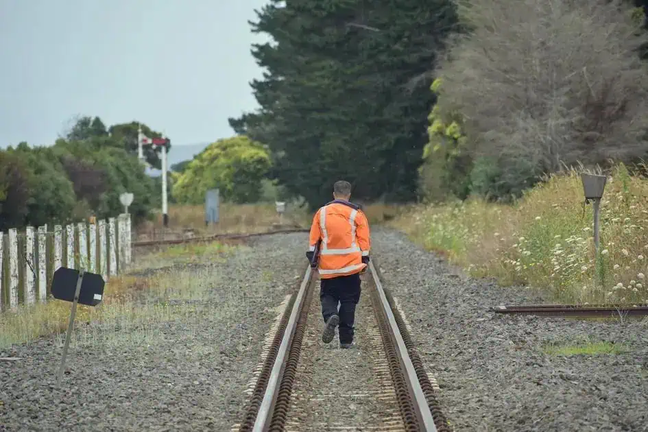 Homem de costas caminhando sobre trilhos de trem em área rural, vestindo jaqueta laranja, representando segurança e prevenção em operações ferroviárias.