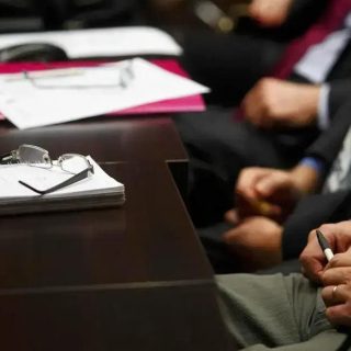 Somentes uma mesa de reunião com documentos, óculos e pessoas de terno presentes em uma audiência ou reunião jurídica.