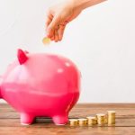 Dinheiro sendo colocado em um cofrinho rosa, simbolizando economia ou investimento, com pilhas de moedas ao redor, conceito de finanças, economia doméstica e planejamento financeiro, relacionado a notícias de direito e economia.
