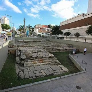 Muros antigos em ruínas no centro de uma cidade brasileira, com pessoas ao redor e edifícios modernos ao fundo, representando conflito entre história e urbanização.