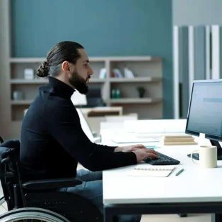 Homem em cadeira de rodas trabalhando em escritório com computador, acessibilidade e inclusão no ambiente de trabalho, direito às facilidades para pessoas com deficiência, direito hoje notícias.