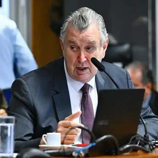A3. Homem de terno falando em comissão durante reunião do tribunal, ambiente formal, com pessoas ao fundo, computadores e microfones, foco em debate jurídico e notícias de direito.