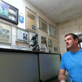 O homem observando uma parede decorada com diversos quadros, fotografias e uma escultura, em ambiente que remete a uma sala de notícias ou escritório de direito.