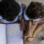 Crianças estudando juntos no chão com caderno e canetas, promovendo educação e inclusão social.