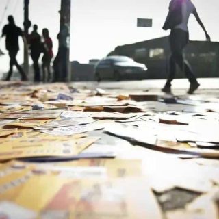Pilhas de jornais e documentos espalhados no chão em uma rua movimentada, pessoas ao fundo, cenário urbano, iluminação de fim de tarde, símbolo de notícias e informação, referente a direito, jornalismo e atualidades no Brasil.