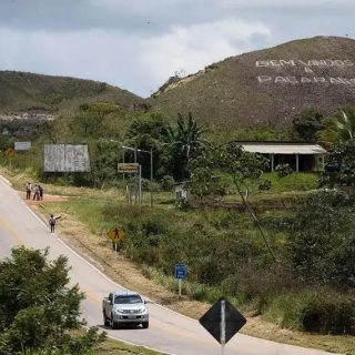 Bandeira de protesto com a frase "Eliminação Padrões" em uma encosta de montanha na zona rural de uma cidade brasileira, cercada por vegetação, com carros e pessoas na estrada, destacando questões ambientais ou sociais.