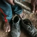Sapatos velhos e usados em solo de terra, mãos sujas de terra e pés descalços, evidenciando trabalho rude ou situação de vulnerabilidade social, tema relevante para notícias de direitos e justiça.